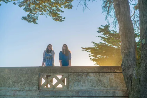 Julie & Emma standing on a bridge with the sun rays shining up in the background